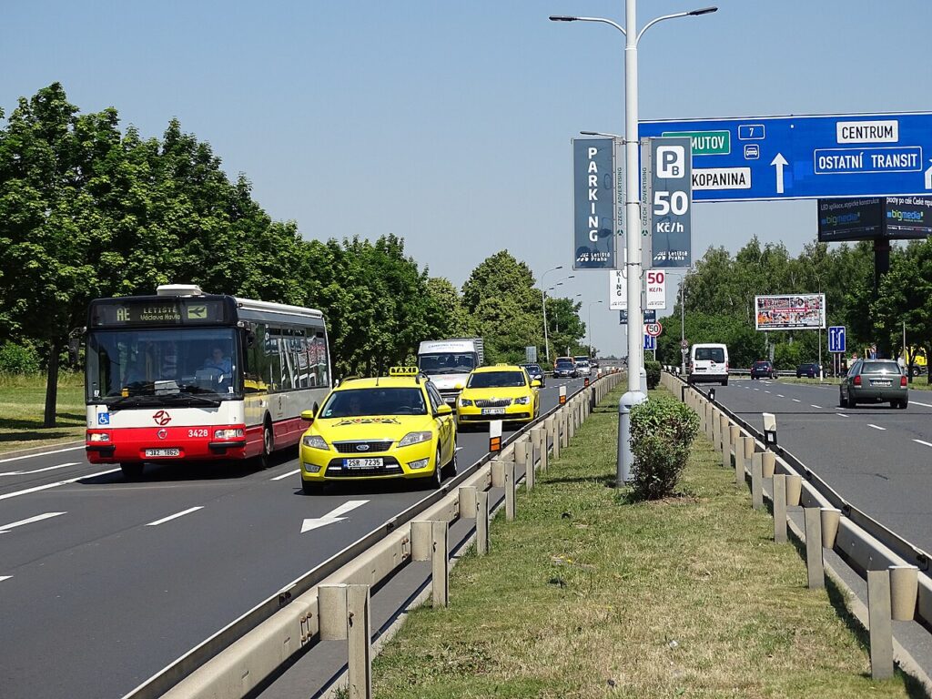 Autobus, taxíky a další vozidla na příjezdové silnici k letišti Praha-Ruzyně, nad vozovkou dopravní značky s nápisy Centrum a Ostatní tranzit